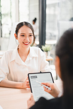 Young woman smiling during job interview with recruiter holding resume on digital tablet in modern office