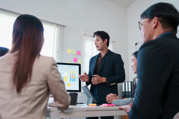 Business team meeting in modern office, young Asian man presenting data on monitor, colleagues listening and discussing strategy, teamwork and collaboration, professional atmosphere