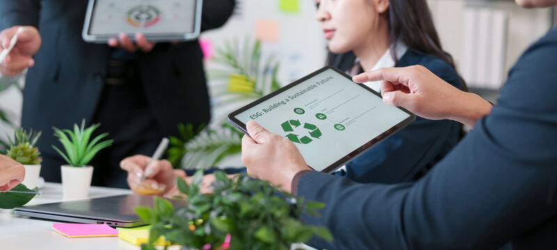 Business team meeting with digital tablet showing sustainability concept, diverse professionals discussing eco friendly strategy, green plants on table, modern office, collaboration, innovation