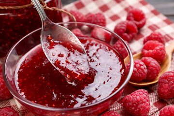 Taking sweet raspberry jam with spoon from glass bowl on table, closeup