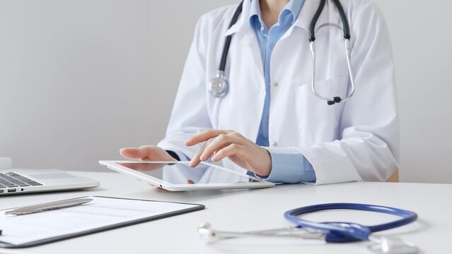 Female doctor wearing white medical coat and stethoscope using digital tablet with laptop and medical records on desk in medical office, showcasing modern healthcare technology. Medicine and helthcare