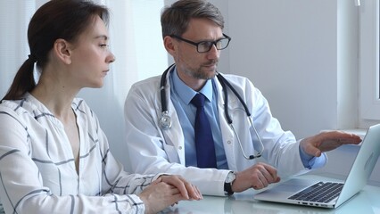 Male doctor showing medical test results on a laptop to a female patient, discussing diagnosis and treatment options in a bright medical office. Medicine and health care
