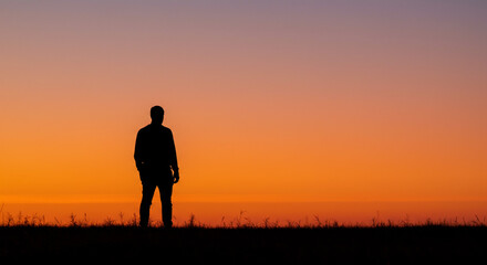 Silhouette of a man standing against a vibrant orange sunset with a dark horizon and grass blades