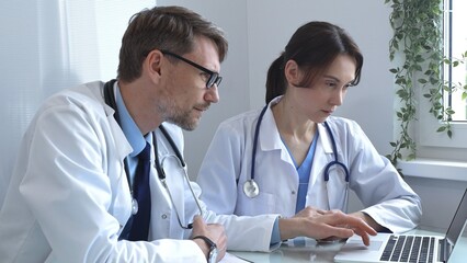 Two doctors are studying medical charts on a laptop, discussing patient data in a hospital or clinical setting. Medicine and healthcare concept.