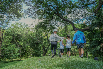 Fototapeta premium A happy Asian family with two young sons enjoys a walk in the park. Parents hold their children's hands while exploring a lush green hill together, a concept of family bonding.