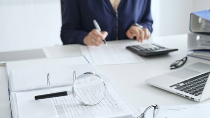 Close up of magnifying glass and calculator over financial documents opposite female accountant analyzing data, reviewing reports, binders and laptop on a white desk. Audit and taxes concept