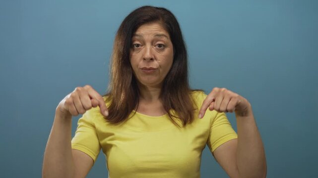 Woman wearing a yellow t-shirt pointing both index fingers down inside a studio against a bright blue wall; serious guidance.
