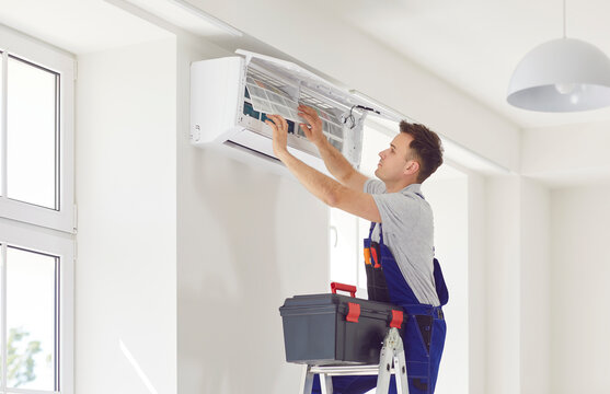 Male worker from AC maintenance service, standing on ladder with toolbox, opens modern white air conditioner on wall, repairs some inside parts and changes filters