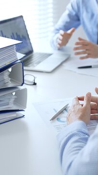 Office workers or accouters are analyzing financial reports and signing documents while sitting at a white table with a stack of blue folders full of paperwork in the foreground. Audit and taxes in