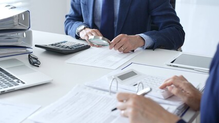 Financial analysts examining documents, using magnifying glass and calculator while studying corporate data at workplace. Audit and taxes in business