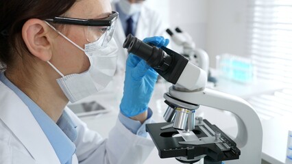 Female scientist wearing mask and gloves using microscope doing scientific research in modern laboratory with colleague working in background. Medicine, healthcare and science concept