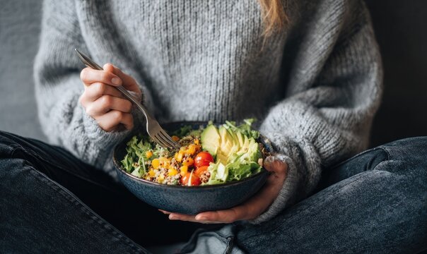 Woman seated, holding a bowl of quinoa salad with avocado, corn, and cherry tomatoes, eating with a fork - Powered by Adobe