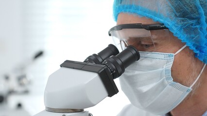Masked scientist examining sample through microscope, wearing protective lab gear during medical research. Medicine, healthcare and science concept