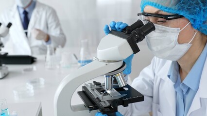 Female scientist wearing a mask and blue safety gear, using a microscope for research while a male colleague works in the background. Medicine, healthcare and science concept