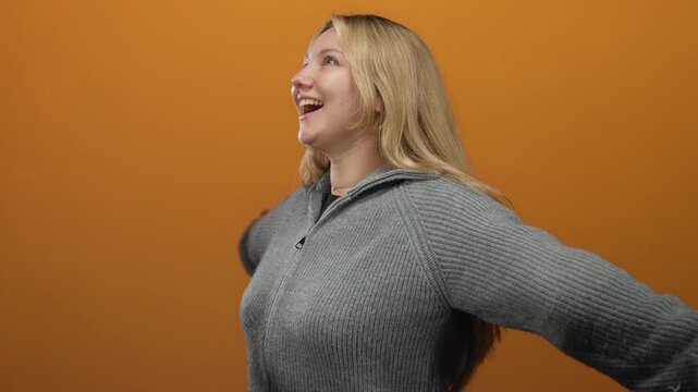 Woman smiling with arms outstretched in front of an orange wall, wearing a cozy grey sweater, conveying a sense of joy and freedom.