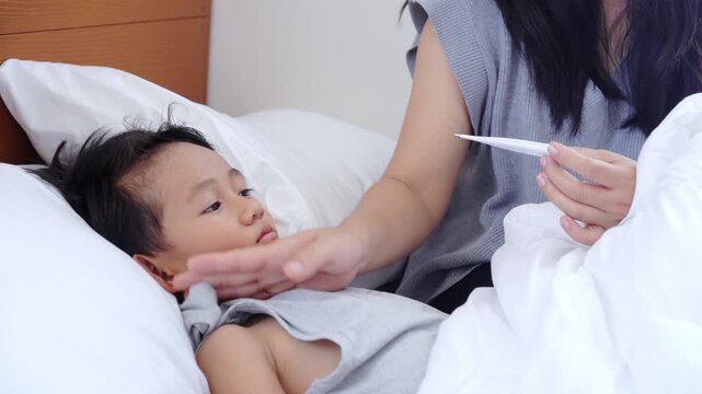 Asian mother checks her sick son's temperature by holding a themometer and touching his forehead The child is lying in bed with a high fever