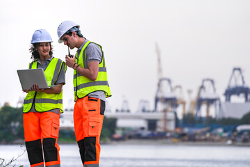 Fototapeta premium Two engineers wearing safety vests and helmets inspect port operations near the waterfront. They use a laptop and walkie-talkie, symbolizing teamwork, logistics, and marine industry work.