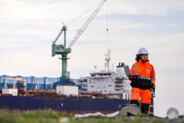 Two port engineers in high-visibility orange uniforms and hard hats communicate while inspecting dockside logistics using a laptop and walkie-talkie, symbolizing teamwork and safety.