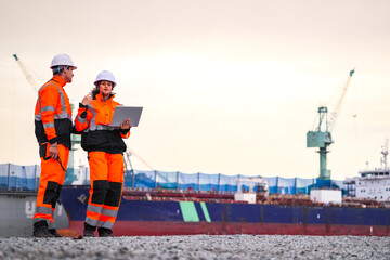 Two port engineers in high-visibility orange uniforms and hard hats communicate while inspecting...