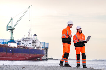 Two engineers wearing high-visibility safety gear and hard hats review plans on a laptop at a shipyard, with a large cargo ship and cranes in the background during sunset.