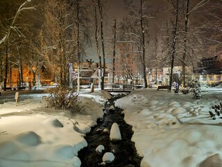 Snow-covered park during evening with a gently winding stream in the center, bordered by trees dusted with snow. Zakopane, Poland 