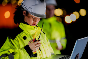 Two diverse engineers coordinate operations on a night shift. The man uses a radio while the woman...