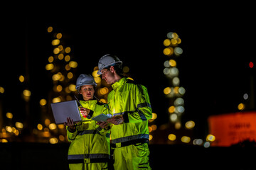 Two diverse engineers coordinate operations on a night shift. The man uses a radio while the woman explains data from a digital tablet, a concept for industrial communication.