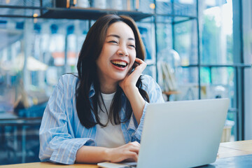 Cheerful Asian woman smiling and laughing while using laptop at modern cafe, representing freelance lifestyle, digital nomad, and online business success.
