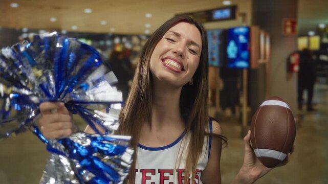 Young hispanic cheerleader holds blue pompom and football while smiling in airport terminal; excitement.