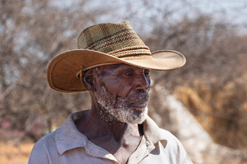 portrait of an old african farmer with a straw hat in the field, dry grass and acacia trees in the bush