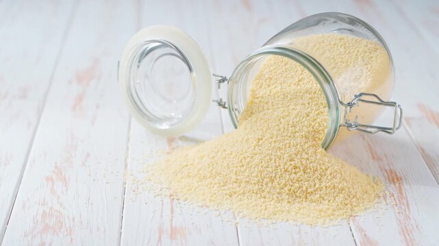 Organic couscous spill out of a glass storage jar a light kitchen table. Raw couscous scattered out of glass jar.
