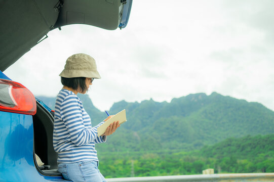 Woman writing in notebook for digital detox. Self-reflection, journaling habit, and mindful planning beside blue car with mountain landscape background. Road trip travel lifestyle. Mindful travel.