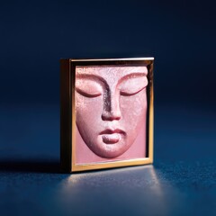 A serene, rosy-hued face sculpted in bas-relief, framed in gold, rests on a dark blue surface.  The piece exhibits subtle textural details and a calm expression