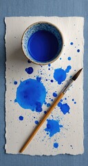 A small bowl of vibrant blue paint sits atop a sheet of textured white paper, splattered with paint droplets, alongside a paintbrush
