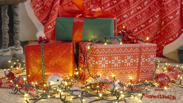 Close-up of Christmas presents in red and green wrapping with bows, festive lights and ornaments on floor with holiday blanket in background