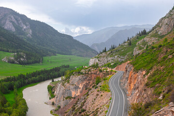 Mountain pass with empty winding Chuysky trakt road along a steep cliff above Chuya river at cloudy summer day in Altai Republic, Russia. Russian route R256 or Chuya Highway at Altai, Siberia.