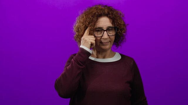 Middle-aged hispanic woman wearing glasses points to head against vibrant purple backdrop, conveying thoughtfulness and creativity.