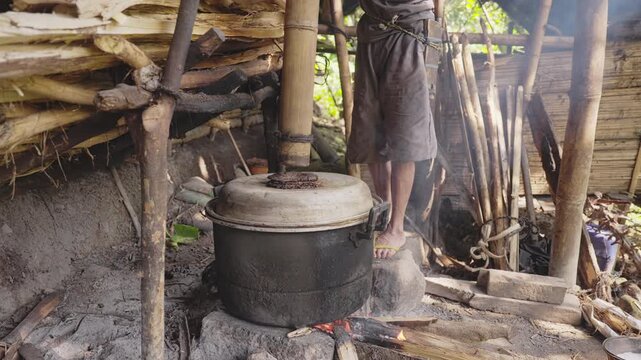 Indonesian palm wine farmer fitting a bamboo pipe on metal pot over open firewood in rural distillery, distilling fermented sap producing arak or sopi alcohol, southeast asia