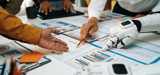 Professionals engage in a collaborative effort, using both human and robotic hands to analyze detailed business charts in a modern office environment, emphasizing innovation.Noogenesis