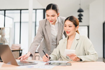 Fototapeta premium Two Asian businesswomen in modern office, one standing and pointing at laptop screen, other sitting and holding calculator, working together on financial report with teamwork and focus