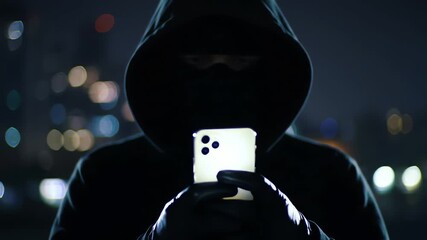 Anonymous hooded hacker in a mask using a smartphone at night with city lights background - Powered by Adobe