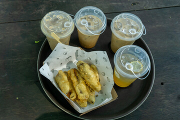 tray with Pisang goreng or banana fritters and four beverages isolated on wooden table. The beverages include three iced coffee drinks and one iced tea or lemonade with a lemon slice.