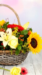 A wicker basket overflowing with a vibrant mix of sunflowers, lilies, roses, and gerbera daisies sits on a rustic whitewashed wooden surface