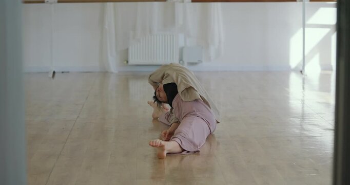 Girl stretches in parterre wide ballet stance while sitting on floor in dance studio. Young woman sits in splits and reaches for her legs during warm-up in studio