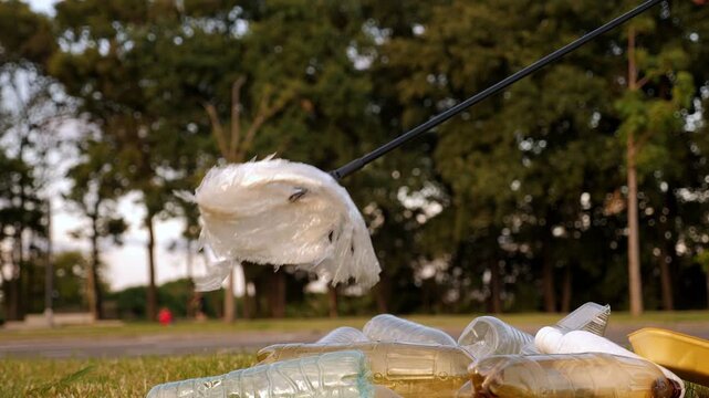 A close-up shot of a woman activist collecting plastic trash with tongs in the green grass. She is helping to save nature from the harmful effects of plastic waste.