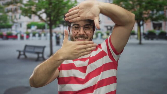 Young man with glasses and beard smiling while framing with hands outdoors on a city street wearing a red striped shirt in a relaxed open space setting.