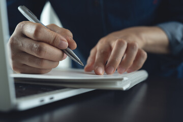 Business woman hand with a pen writing on paper notebook with laptop on office table, planning work project, closeup.  business plan, job schedule,