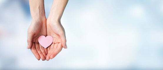 A pair of human hands gently holding a pink heart shape against a soft blue background. Symbolizing...
