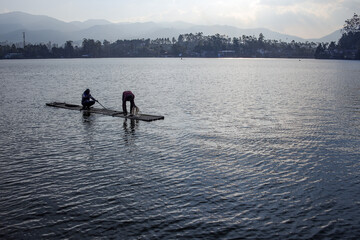 Fototapeta premium Two Fishermen On A Bamboo Raft, Preparing Their Nets On The Lake, With Mountains In The Background