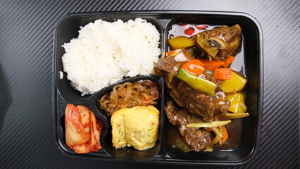 Top view of a Korean-style bento meal featuring white rice, braised beef with vegetables, kimchi, japchae, and rolled omelette.
